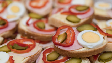 Open-face sandwiches with sausage and vegetables as a background. Close-up of white bread slices with ham, cheese, hard-boiled eggs, capsicum and cucumber. Small depth of sharpness.