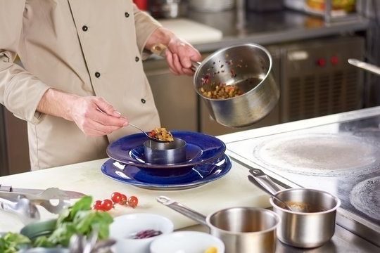 Chef hands putting vegetables ratatouille in plate. Male chef at professional kitchen holding spoon and saucepan with stewed vegetables.