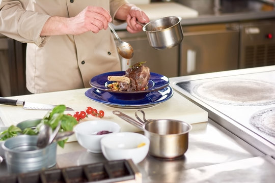 Chef pouring with sause lamb shank. Male chef hands at professional european kitchen pouring lamb shanks with sauce. Tasty and nutrition dish.