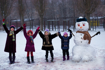 A small cheerful girl near big funny snowman. A cute little girl has fun in winter park, wintertime