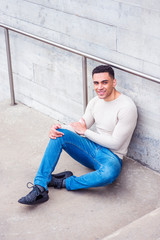 Young handsome American Man wearing light gray knit sweater, blue jeans, black sneakers, sitting on ground by wall in corner of street in New York, smiling, relaxing..