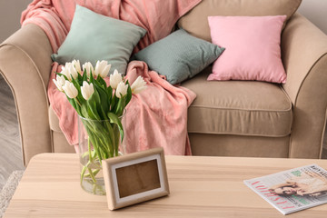 Wooden table with bouquet of tulips and photo frame near sofa in living room
