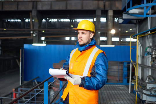 Confident bearded technician wearing reflective vest and protective helmet standing at spacious production department of modern plant and taking notes while carrying out inventory