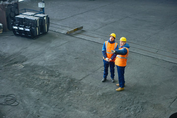 High angle view of two engineers wearing hardhats and uniform standing at spacious production department of modern plant and brainstorming on faced issue