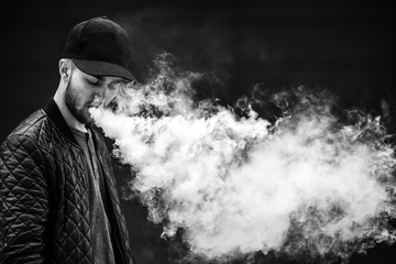 Vape man. Portrait of a handsome young white guy in a modern black cap vaping and letting off puffs of steam from an electronic cigarette opposite the
