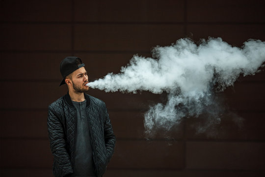 Vape man. Portrait of a handsome young white guy in a modern black cap vaping and letting off puffs of steam from an electronic cigarette opposite the