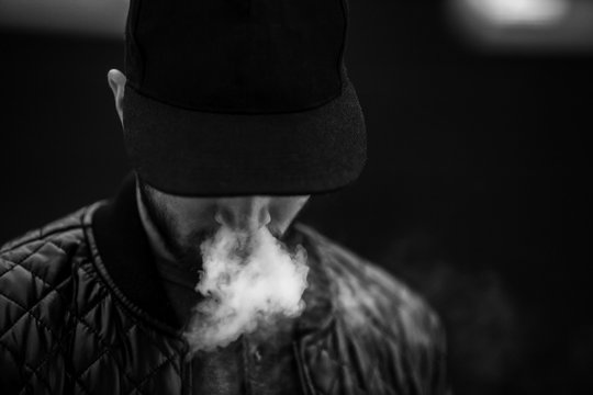 Vape man. Portrait of a handsome young white guy in a modern black cap vaping and letting off puffs of steam from an electronic cigarette opposite the