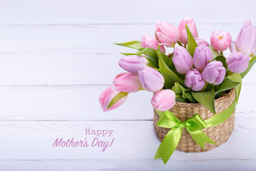 Bouquete of pink tulips in a wicker basket on white wooden background, with text Happy mother's day
