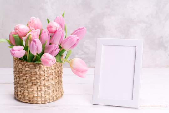 Bouquet of pink tulips in a wicker basket and empty picure frame on light gray background