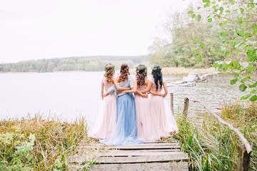 in full growth three bridesmaids in powdery dresses transformers and wreaths on the head embrace the bride in a blue dress on a wooden bridge and look at the water - obrazy, fototapety, plakaty