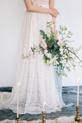 a wedding spring bouquet from Ranunculus in the hands of the bride in a lacy white dress against a white wall and burning candles