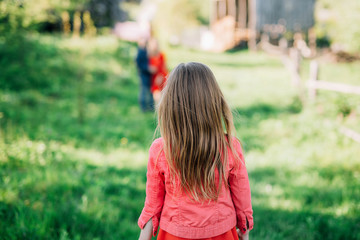 a girl in a red dress and a jacket with flowing hair will look at parents who stand in the distance on a green meadow