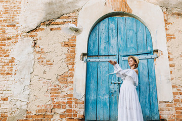 pregnant girl in a white long dress and a straw hat launches a dove and laughs against a blue door and a brick wall background