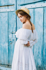 Fototapeta premium portrait of a pregnant girl in a white long dress and a straw hat that behind her back has brought her hands against the blue door