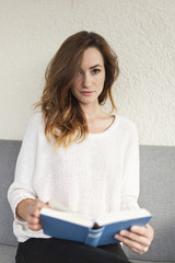student women on a couch in white cardigan holding a book focusing the viewer