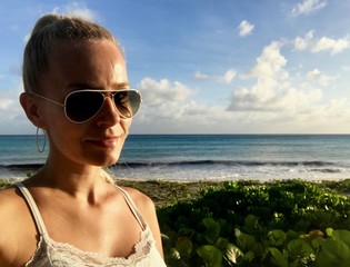 Beautiful blonde female model with sunglasses in Oistins, Barbados at a untouched beach with lush greenery, crystal clear turquoise water and a blue sky with white clouds