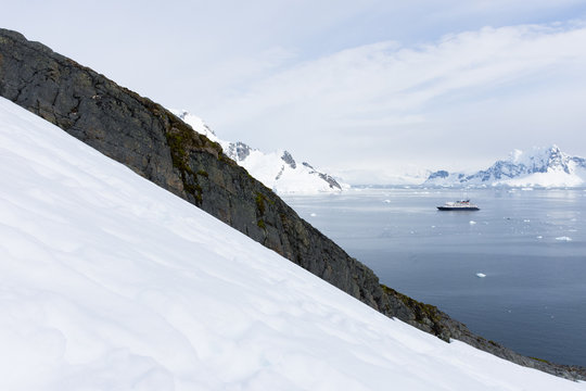 Ship seen in Paradise Bay viewed from the Antarctic Peninsula. Snow and a ridge of rocks are seen in the foreground.