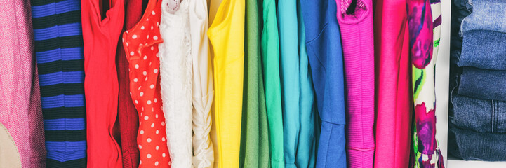 Clothing store clothes hanging in shopping rack. Banner texture background of different selection of fabrics and patterns. Panoramic crop. Shelves of colorful rainbow assorted outfits.