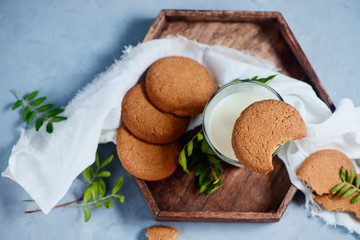 Fototapeta na wymiar Cookies with bite marks, a glass of milk, wooden tray and tiny green leaves decor on a stone background with white copy space. Healthy breakfast concept from above.