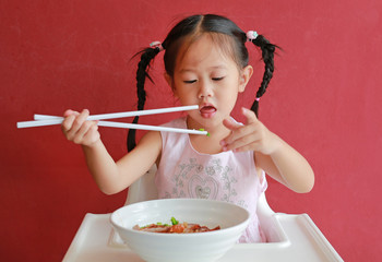 Little asian girl eating Egg noodle with roasted duck and roast pork on high chair against red wall background.
