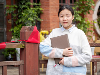 Portrait of a young beautiful Chinese girl hold a notebook and smile at camera, against blur school...