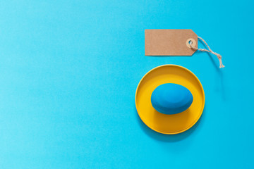 A blue Easter egg is lying in a brightly yellow metal box. Near the empty space on the craft label with your text. Blue background. Copy space.