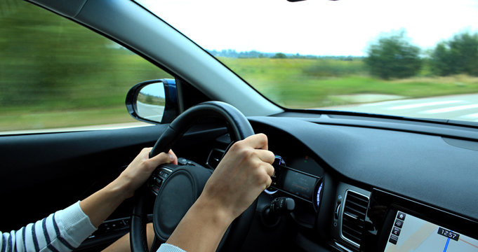 The driver's hands lie on the steering wheel of a car at speed on a country road
