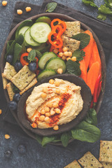 Homemade traditional hummus with vegetables, crackers on a black clay dish, dark background, top view. Healthy vegan food concept.
