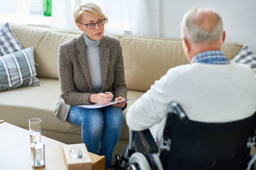 Portrait of mature female psychiatrist interviewing handicapped senior man during therapy session,...