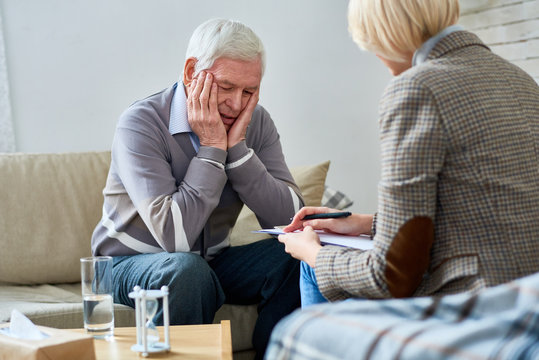 Portrait of depressed senior man sharing problems during therapy session with female psychiatrist writing on clipboard