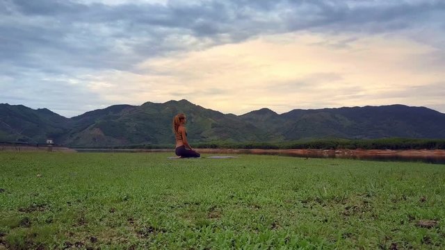 Flycam Removes from Girl Sitting in Yoga Pose on Lake Shore