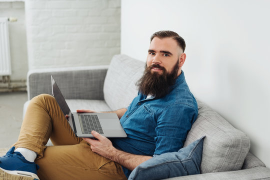 Smiling bearded man sitting on sofa with laptop