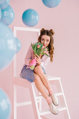 A girl in a blue dress and a pink shirt sits on a ladder in a pink room with a bouquet of tulips