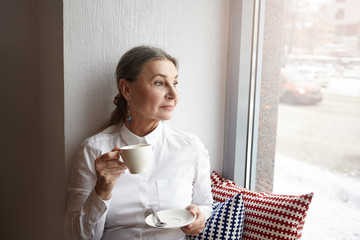 Beautiful middle aged female with gray hair and blue eyes sitting at cafeteria on windowsill, enjoying morning coffee, holding cup and looking through window, havign thoughful facial expression