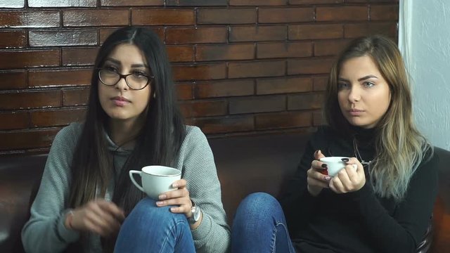 Two yong girls drinks coffee and talking sitting on coush.