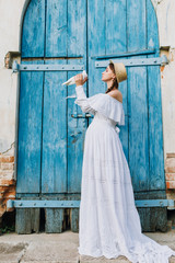 pregnant girl in a white long dress and a straw hat is holding a pigeon in her hands and wants to kiss him against a blue door and a brick wall