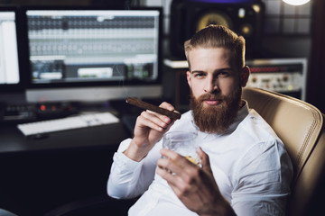The male producer sits in the recording studio behind the remote control and smokes a cigar.