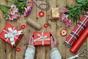 Festive background. Top view composition of woman hands wrap present for Birthday, Mother's day, Valentine's Day, March 8. Packed gifts and flowers, wrapping paper and tools on shabby wooden table