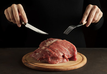Hands holding fork and knife and ready to eat raw pork meat on the wooden cutting board on dark...