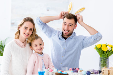 Happy family playing with bunny ears by table with colored eggs for Easter