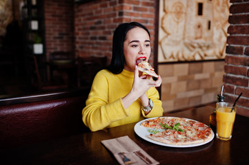 Funny brunette girl in yellow sweater eating pizza at restaurant.