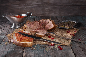 Baked succulent pork meat, sliced pieces on parchment paper, close-up with bread and tomato sauce, dark wooden rustic background