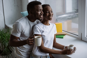 happy young african american couple holding cups of coffee and looking at window