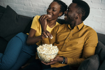 happy young african american couple eating popcorn while sitting on sofa at home
