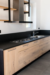 New kitchen in oak and black granite and stainless steel with island, sink, doors, shelves and extractor hood. Industrial design cuisine in a renovated apartment with gray concrete flooring. Cookery