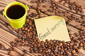 Full green metal bucket of black coffee near paper sticker with inscription good morning and scattered lot of roasted beans on old worn wooden table