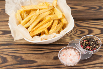 Tasty sticks of french fries in white ceramic bowl with crumpled paper and salt with pepper on old brown rustic wooden table