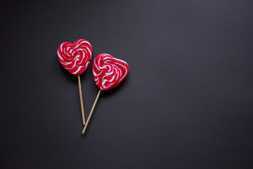 Two red candies on a stick in the shape of a heart lie on the left on a black background. Love concept. Closeup.