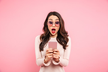 Surprised woman with red lips typing text message or scrolling social network using smartphone, over pink background