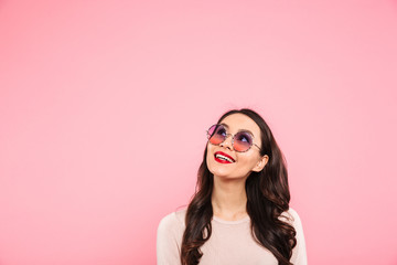 Image of pleased happy adult girl with red lips wearing round sunglasses looking upward on copy space, isolated over pink background